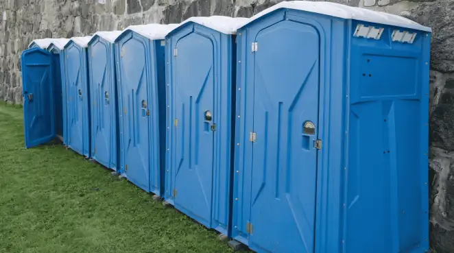 Row of blue portable toilets lined up on grass against a stone wall for event sanitation in Pueblo, CO